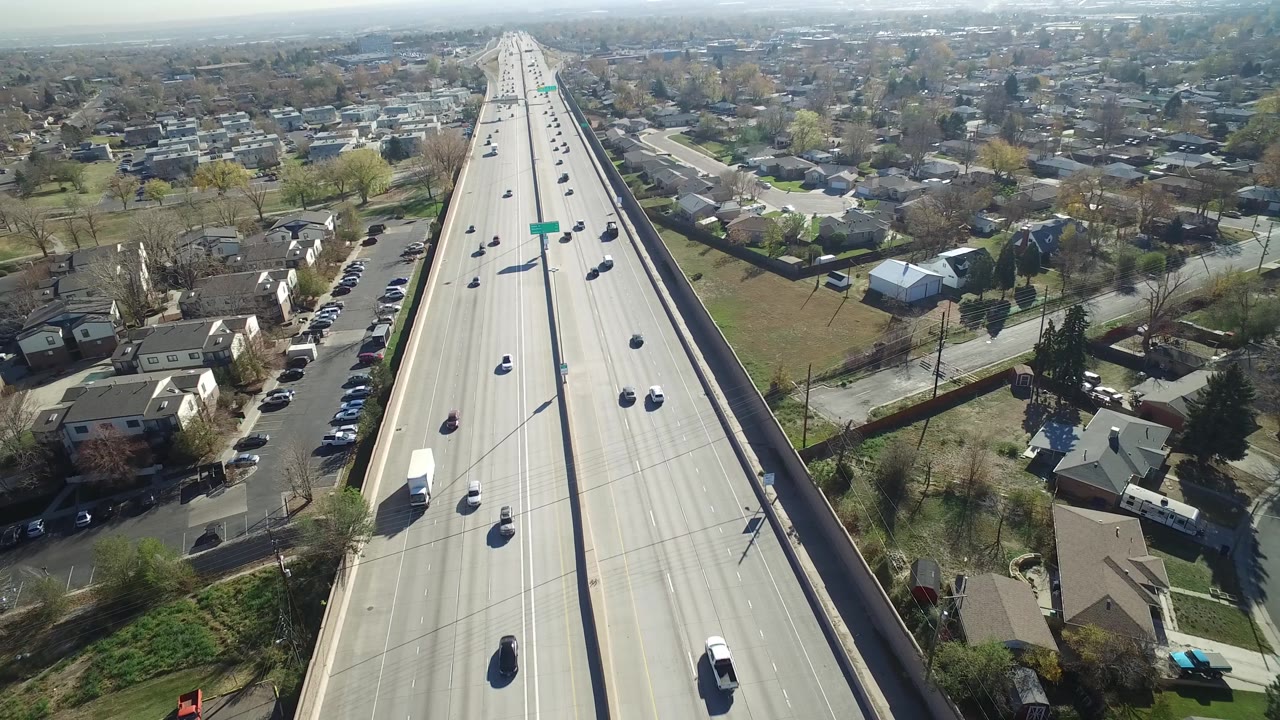 The drone view in Hidden Lake, Colorado looks along the freeway lanes as cars pass between residential blocks, bare trees, green lawns, and no visible people.