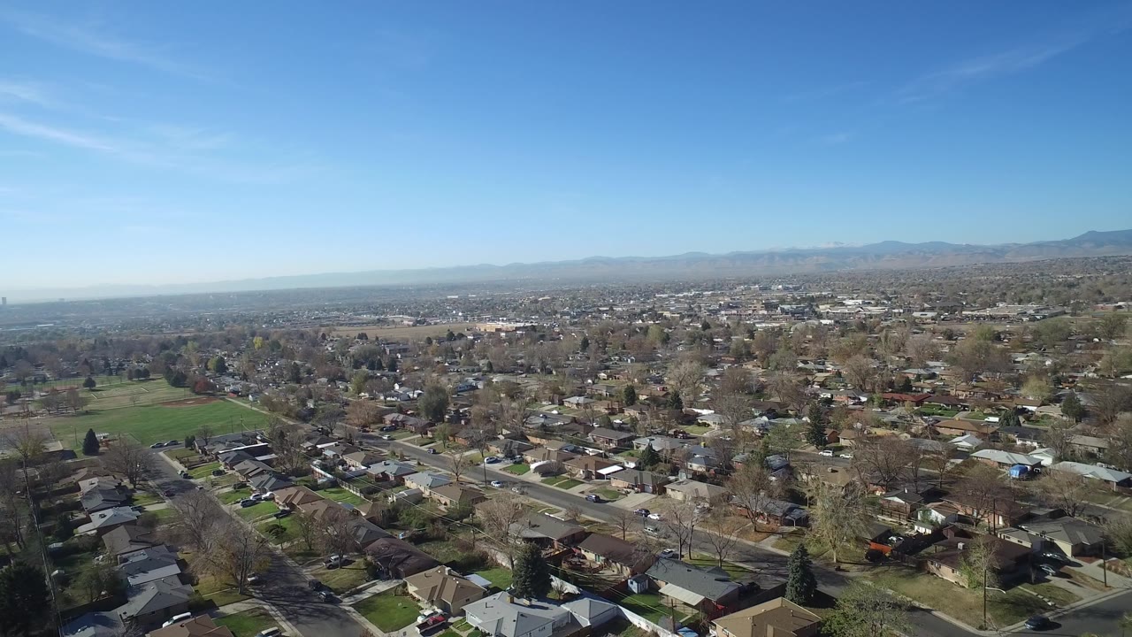 Wide aerial view over Hidden Lake, Colorado homes, clean roads, trees, a green field to the left, distant mountains, and no visible people.