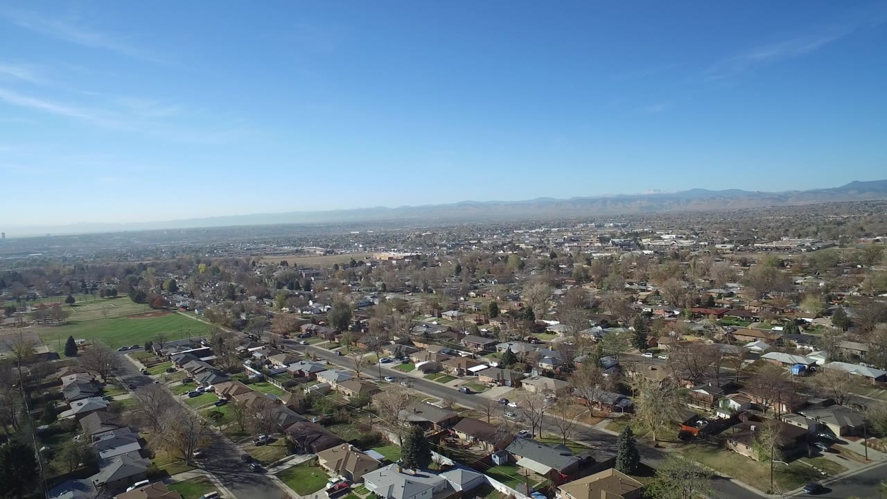 Aerial frame over Hidden Lake, Colorado showing more of the neighborhood grid, a green field at left, scattered trees, distant mountains, and no visible people.