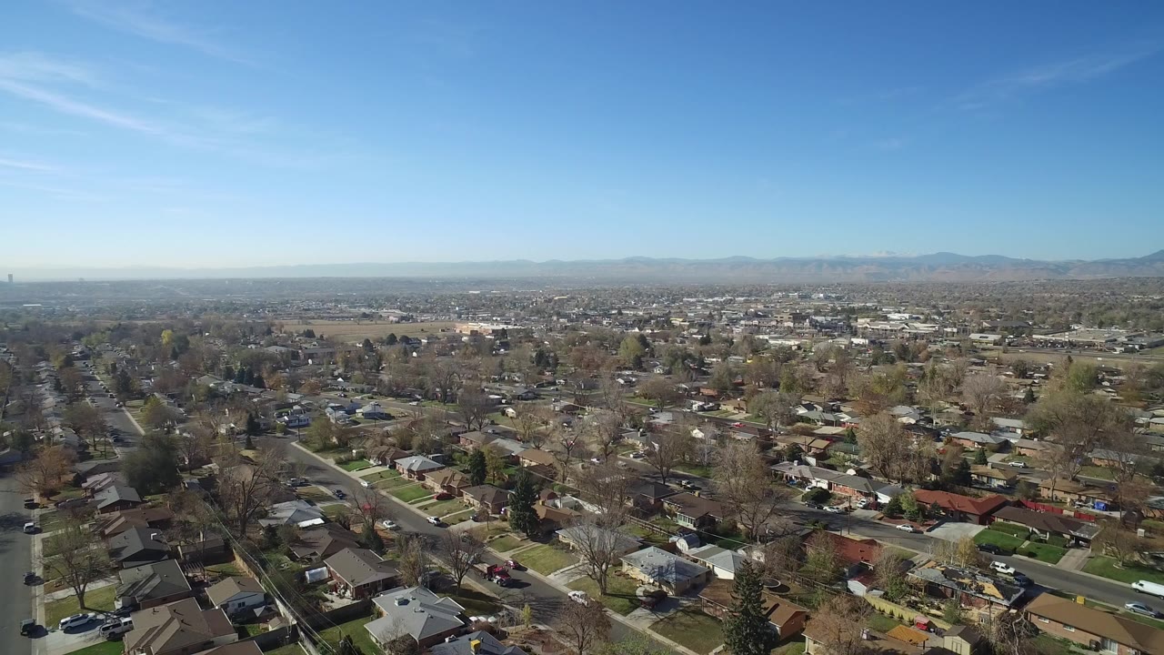 Wide aerial frame over Hidden Lake, Colorado homes and tree-lined streets with distant mountains under clear blue sky, no visible people.