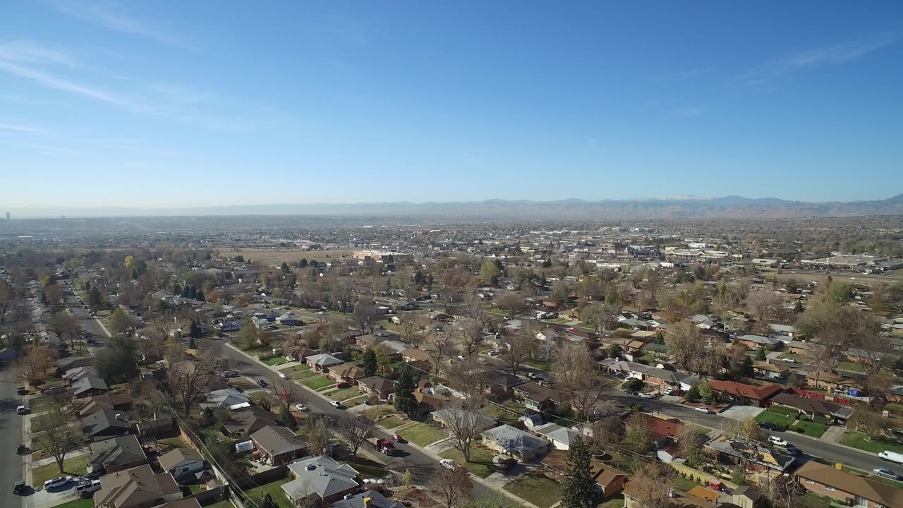 High view across Hidden Lake, Colorado rooftops and neighborhood blocks, with a long road at left and hazy mountains in the background, no visible people.