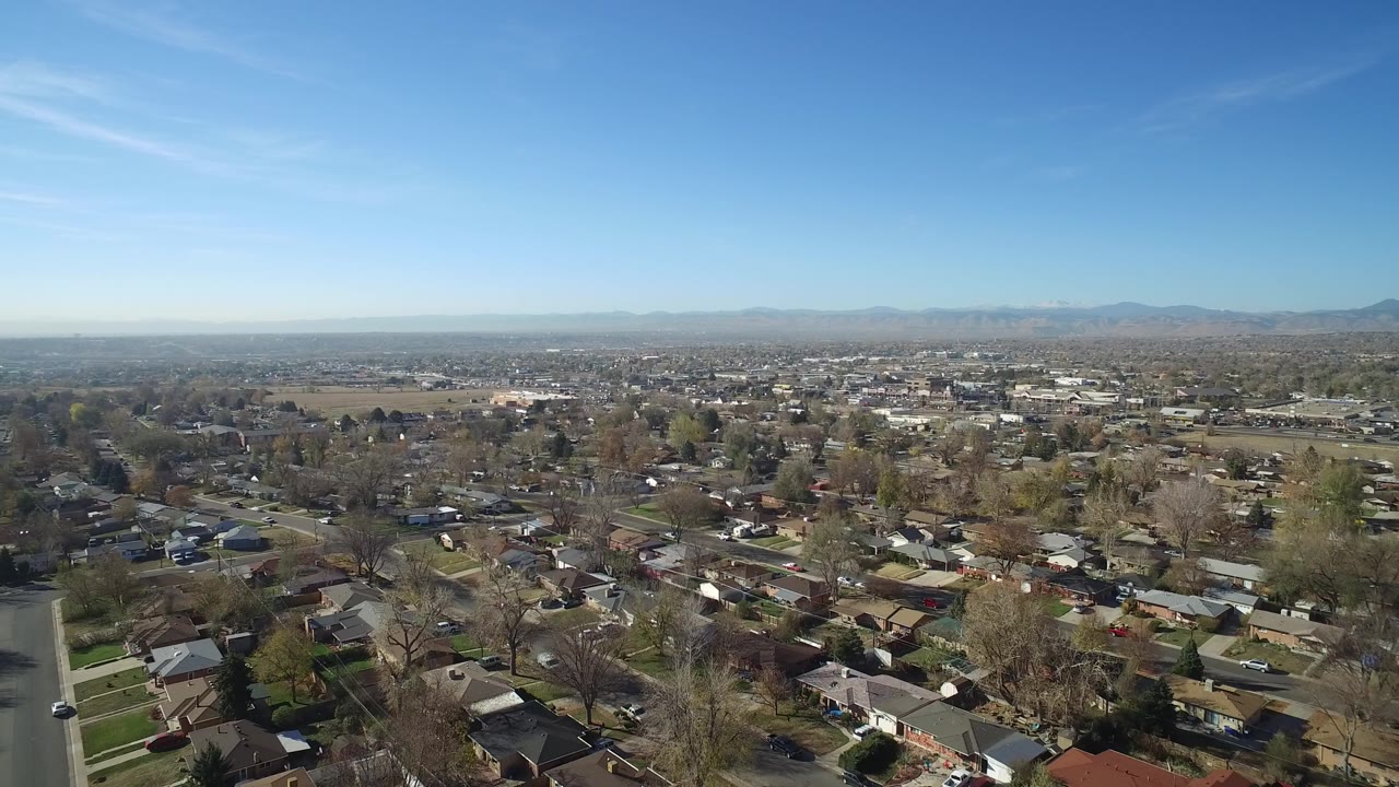 Aerial opening view over Hidden Lake, Colorado neighborhoods with rooftops, tree-lined streets, open land beyond the homes, distant mountains, and no visible people.