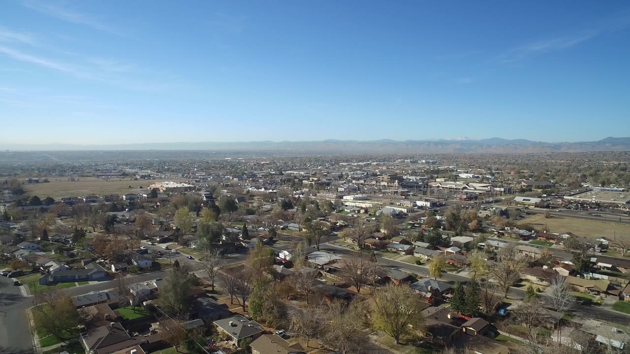 Wide aerial frame over Hidden Lake, Colorado homes and curved residential streets, with winter-bare trees, distant commercial buildings, mountains on the horizon, blue sky, and no visible people.
