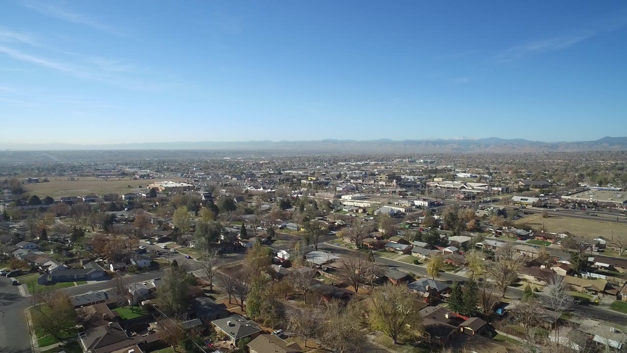Aerial view across Hidden Lake, Colorado with rooftops and streets in the foreground, cars on neighborhood roads, open land and low buildings beyond, distant mountains, and no visible people.