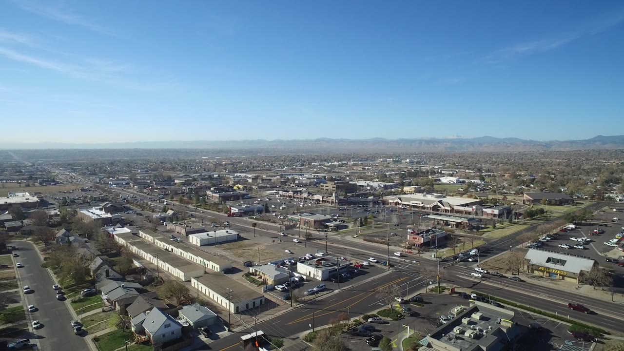 Opening aerial frame over Hidden Lake, Colorado, with a multi-lane road, low commercial buildings, homes, parking lots, distant mountains, and no visible people.
