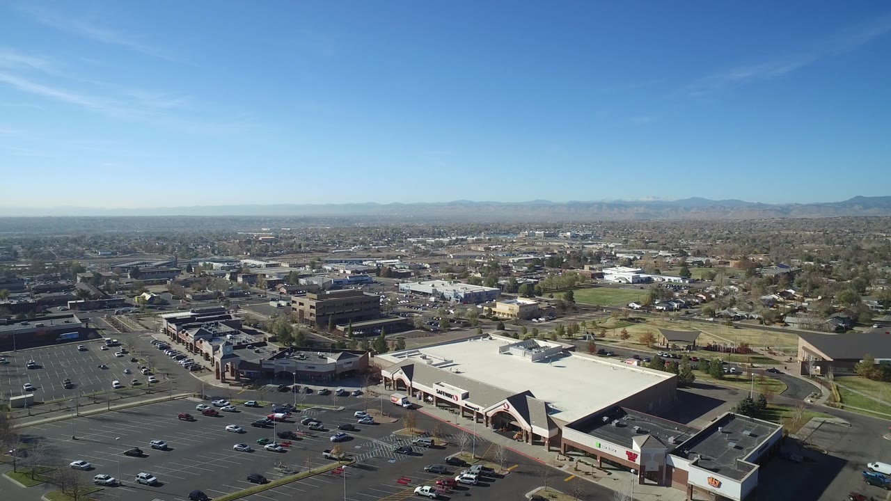 Drone-height view in Hidden Lake, Colorado with the outlet mall and parking rows low in frame, commercial buildings and residential streets across the middle, distant mountains, and no visible people.