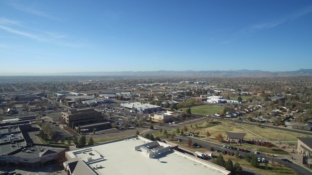 Aerial view over rooftops, roads, trees, parked cars, and distant mountains under blue sky.