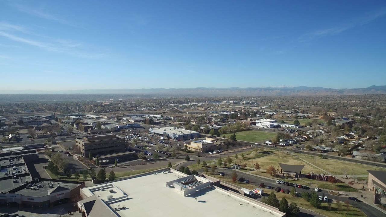 Aerial view in Hidden Lake, Colorado, showing a dark office building at left, pale rooftops, parking lots, open grass, residential blocks, distant mountains, and no visible people.