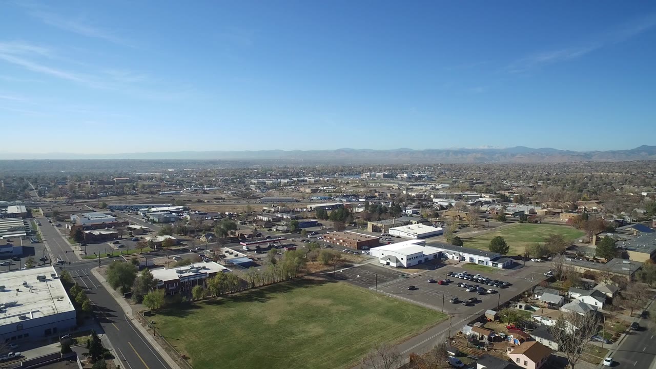 Opening aerial frame over Hidden Lake, Colorado with a road at left, green fields, low white-roofed buildings, distant mountains, clear blue sky, and no visible people.