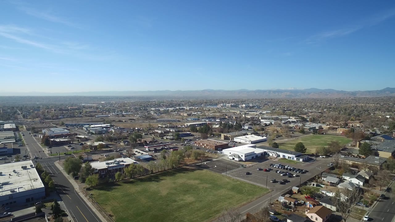 Second aerial frame over Hidden Lake, Colorado showing more of the road corridor, parking areas, white buildings, green lawns, distant mountains, and no visible people.