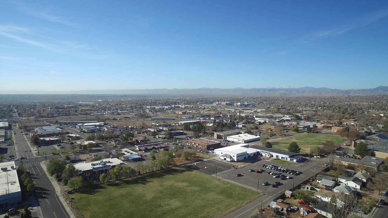 Wide drone view of parking lots, white buildings, green fields, homes, and distant hills.