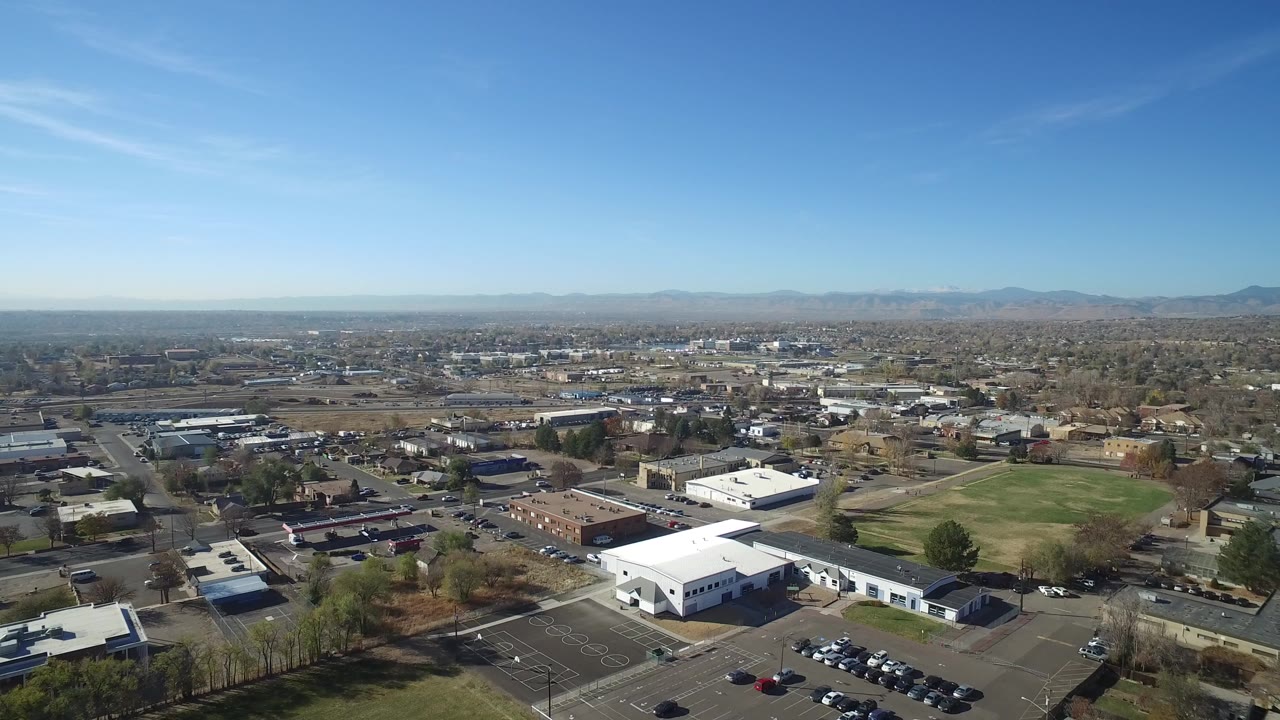 Opening aerial frame over Hidden Lake, Colorado showing white-roofed buildings, a parking lot, a grass field, residential streets, distant mountains, and no visible people.