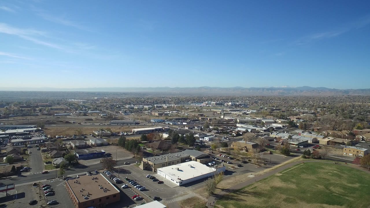 Opening aerial frame over Hidden Lake, Colorado, with a green field and low commercial buildings in the foreground, town blocks beyond, distant mountains, and no visible people.