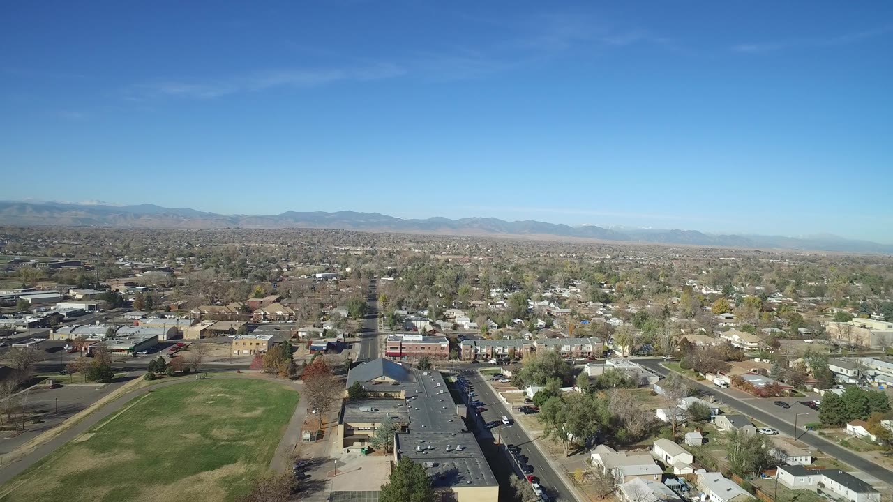 Aerial frame over Hidden Lake, Colorado showing homes around a road grid, a dark parking lot, a rectangular grass field, mountain ridges, and no visible people.