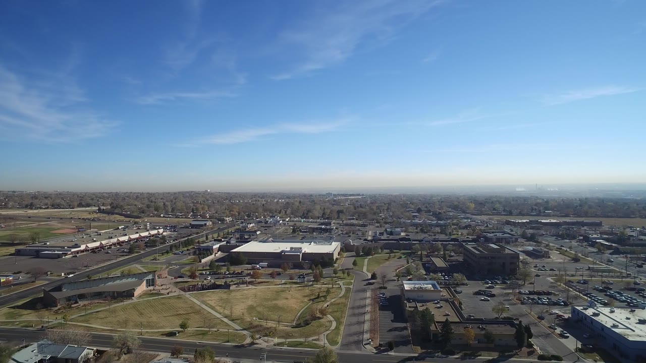 Opening aerial frame over Hidden Lake, Colorado showing roads, parking lots, low commercial buildings, open grass, distant neighborhoods, and no visible people.