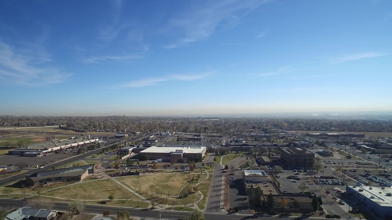 Second aerial frame in Hidden Lake, Colorado with a large flat-roofed building near center, parking lots and roads below, distant neighborhoods, and no visible people.
