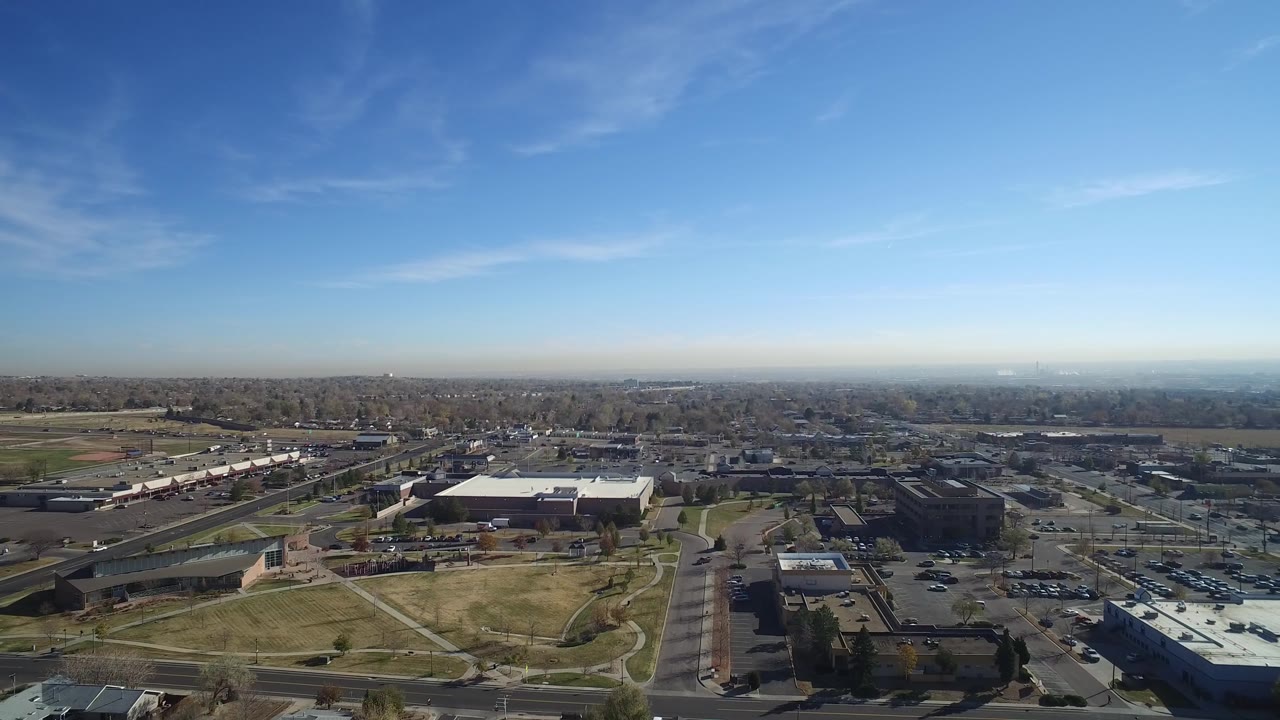 Aerial suburban town view with a broad road, parking areas, low buildings, grass, and a hazy horizon.