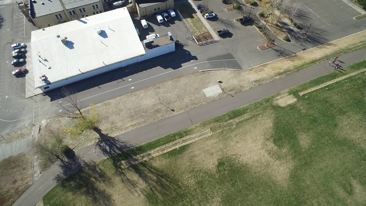 Drone frame over Hidden Lake, Colorado with the white commercial roof near the upper left, parking stalls and cars along the top, open lawn across the lower half, and no visible people.