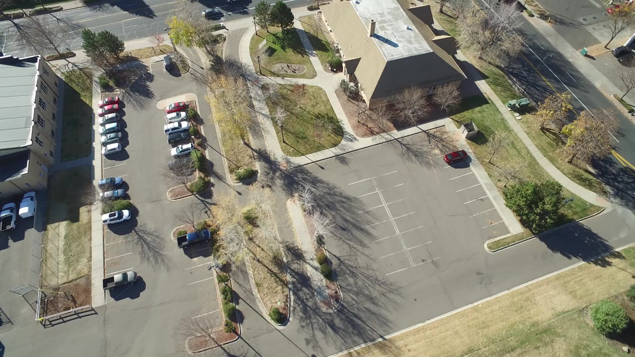 Overhead view of a parking lot, sidewalks, buildings, trees, and nearby roads.