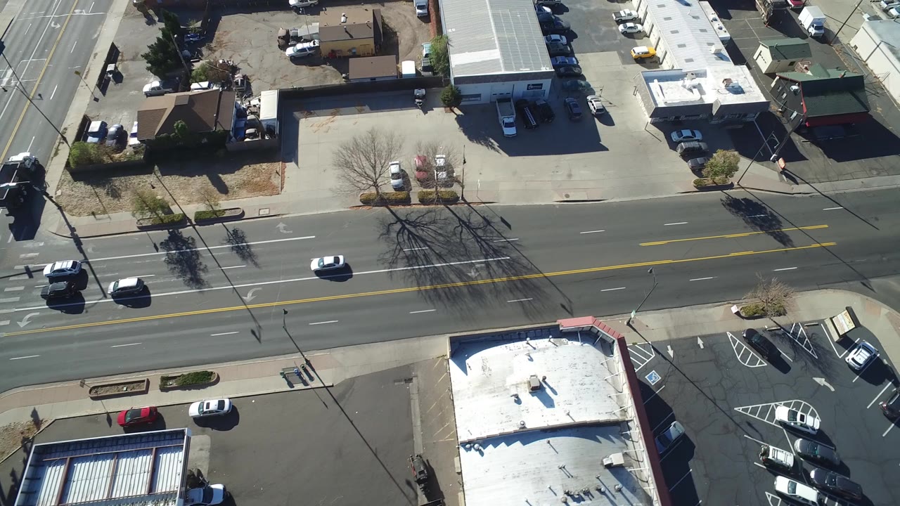 Aerial frame from Hidden Lake, Colorado showing a broad road, parked cars, flat-roofed buildings, bare trees, and no visible people at the start of the clip.