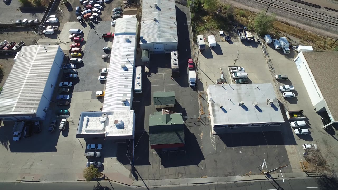Aerial frame in Hidden Lake, Colorado with white-roof commercial structures, rows of parked cars, a small green-roof building, and railroad tracks in the background, with no visible people.