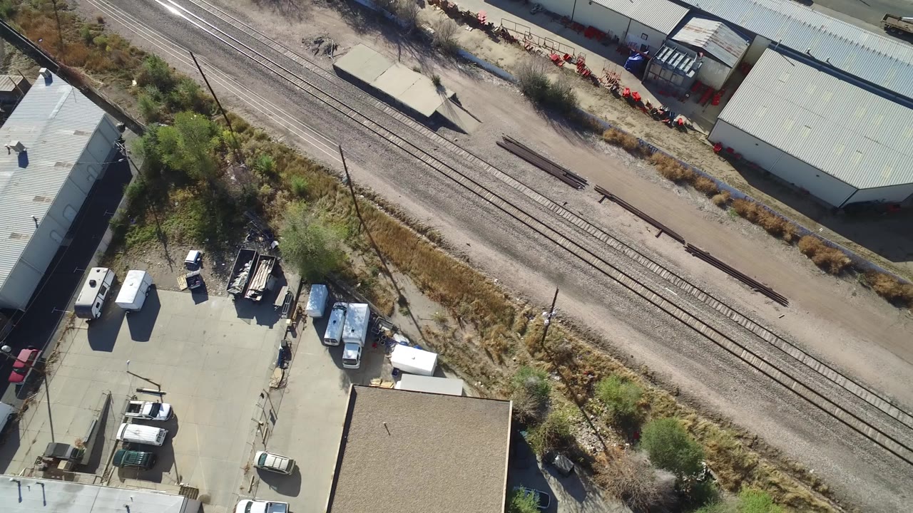Aerial frame over Hidden Lake, Colorado, with rail tracks running diagonally past dry brush, light industrial roofs, parked utility vehicles, and no visible people.