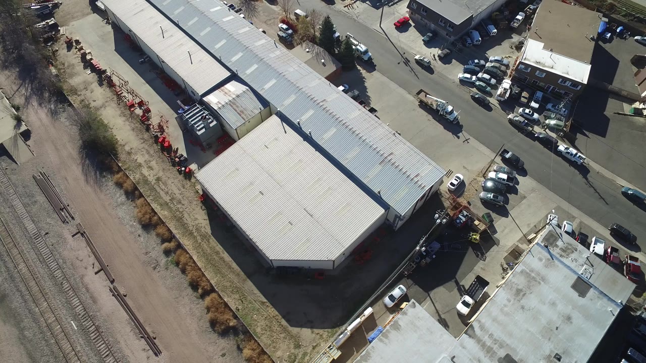 High-angle aerial view of a long warehouse, parking lots, street, and rail tracks.