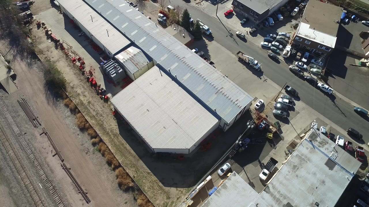 Hidden Lake, Colorado aerial frame with the warehouse roof centered, rows of parked vehicles around nearby buildings, rail tracks at the edge, and no visible people.