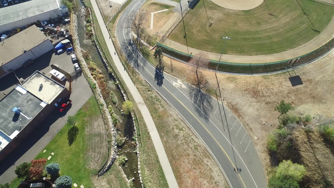 High aerial frame over Hidden Lake, Colorado showing a curved road, sidewalk, green field fence, dry grass, landscaped drainage, parked vehicles, nearby buildings, and no visible people.