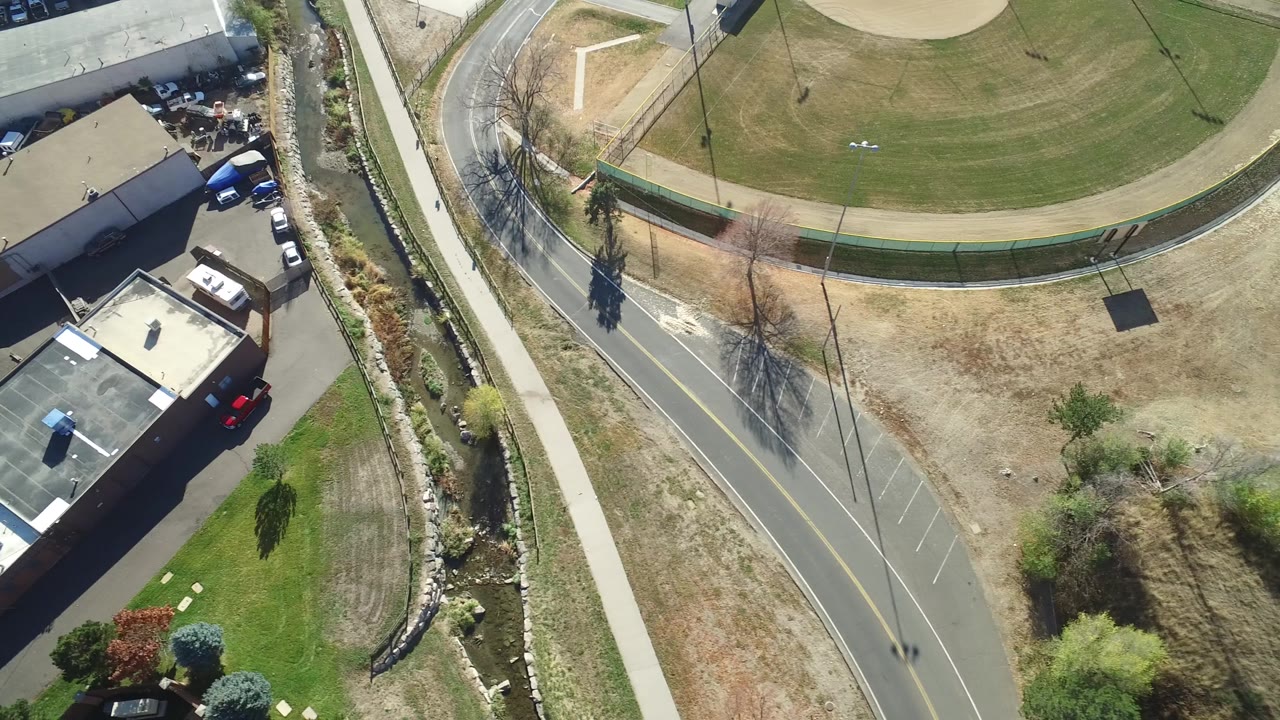 Second aerial moment over Hidden Lake, Colorado with the road bend lower in frame, a sidewalk running beside a planted channel, a fenced green field at top, building edges at left, and no visible people.