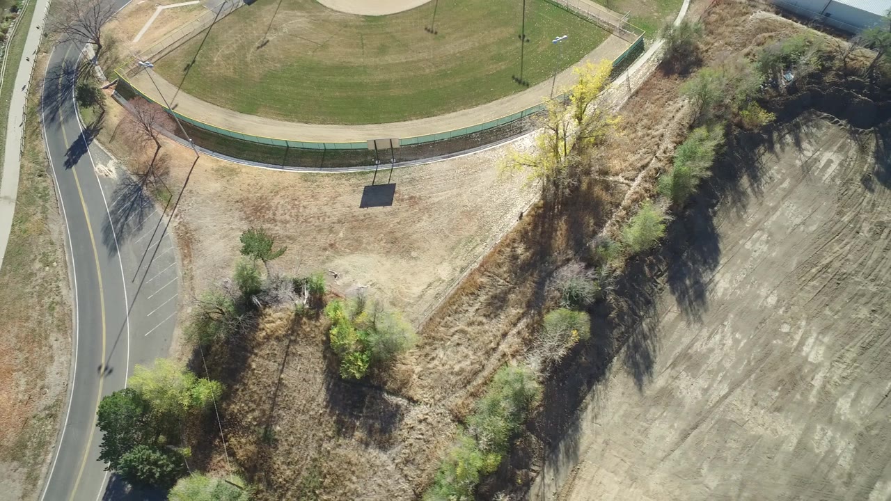 Aerial frame over Hidden Lake, Colorado with the green fenced lawn across the top, a road along the left edge, brush and small trees in the middle, graded dirt field to the right, and no visible people.