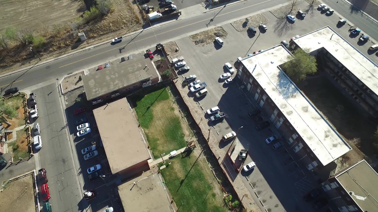 Drone-style overhead frame in Hidden Lake, Colorado showing a long light-roofed residential building, rows of parked cars, nearby roads, small buildings, grass areas, and no visible people.