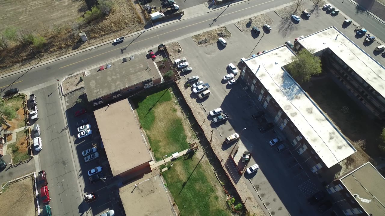 High aerial frame in Hidden Lake, Colorado with a full-looking parking area beside rectangular residential buildings, intersecting streets at the top and left, grass patches, and no visible people.