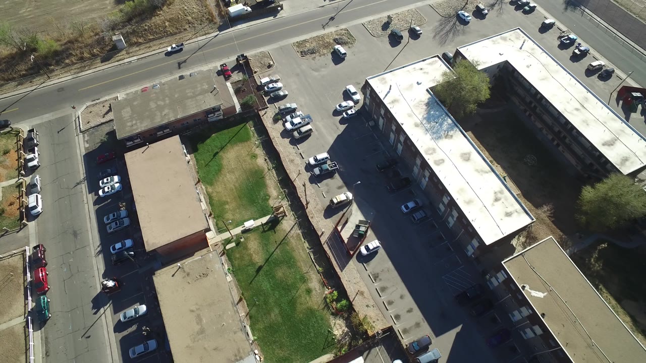 Overhead frame in Hidden Lake, Colorado showing apartment-style buildings, a green lawn strip, cars parked around the lots and along the street, dry ground beyond the road, and no visible people.