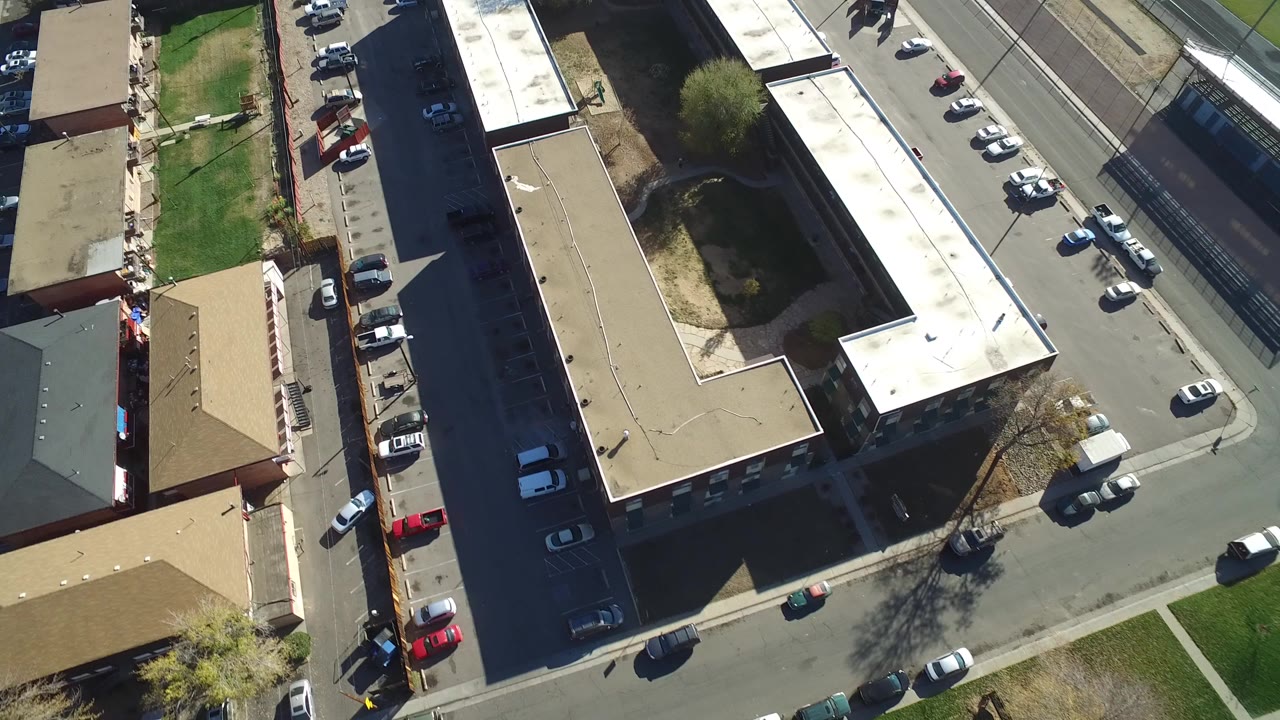 High aerial frame over Hidden Lake, Colorado showing apartment-style rooftops, a street corner, parked cars along curbs and lots, green lawns, and no visible people.