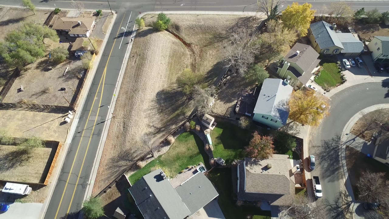 Aerial frame over Hidden Lake, Colorado with the two-lane road near center, residential roofs to the right, pale yards, trees, parked cars, and no visible people.