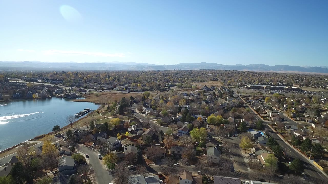 Aerial frame over Hidden Lake, Colorado, showing the lake edge, a bright wake on the water, curving neighborhood streets, clustered homes, and no visible people.