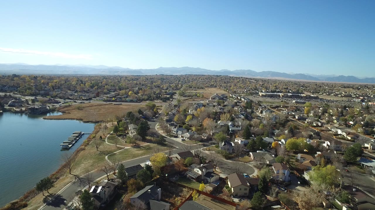 Wide aerial frame over Hidden Lake, Colorado with the lake and docks at left, residential streets and houses below, a mountain horizon in the distance, and no visible people.