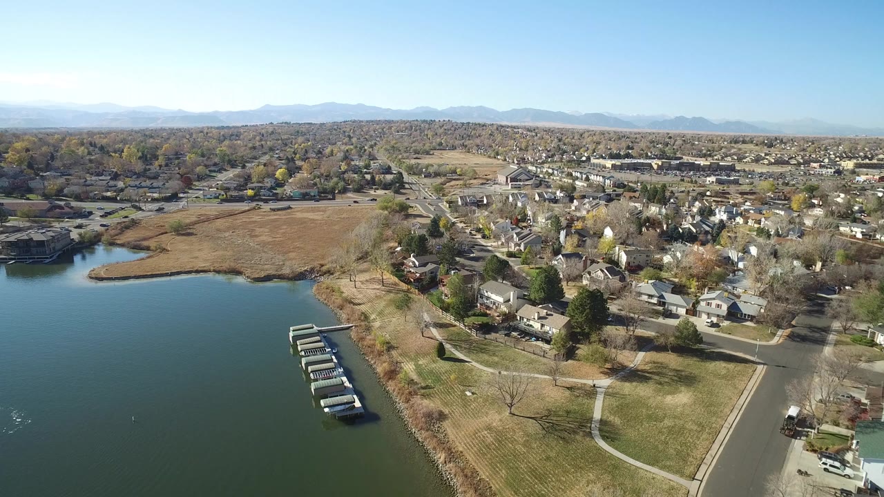 High aerial frame over Hidden Lake, Colorado showing lake water at left, a line of docks, open grass, nearby homes, distant mountains, and no visible people.