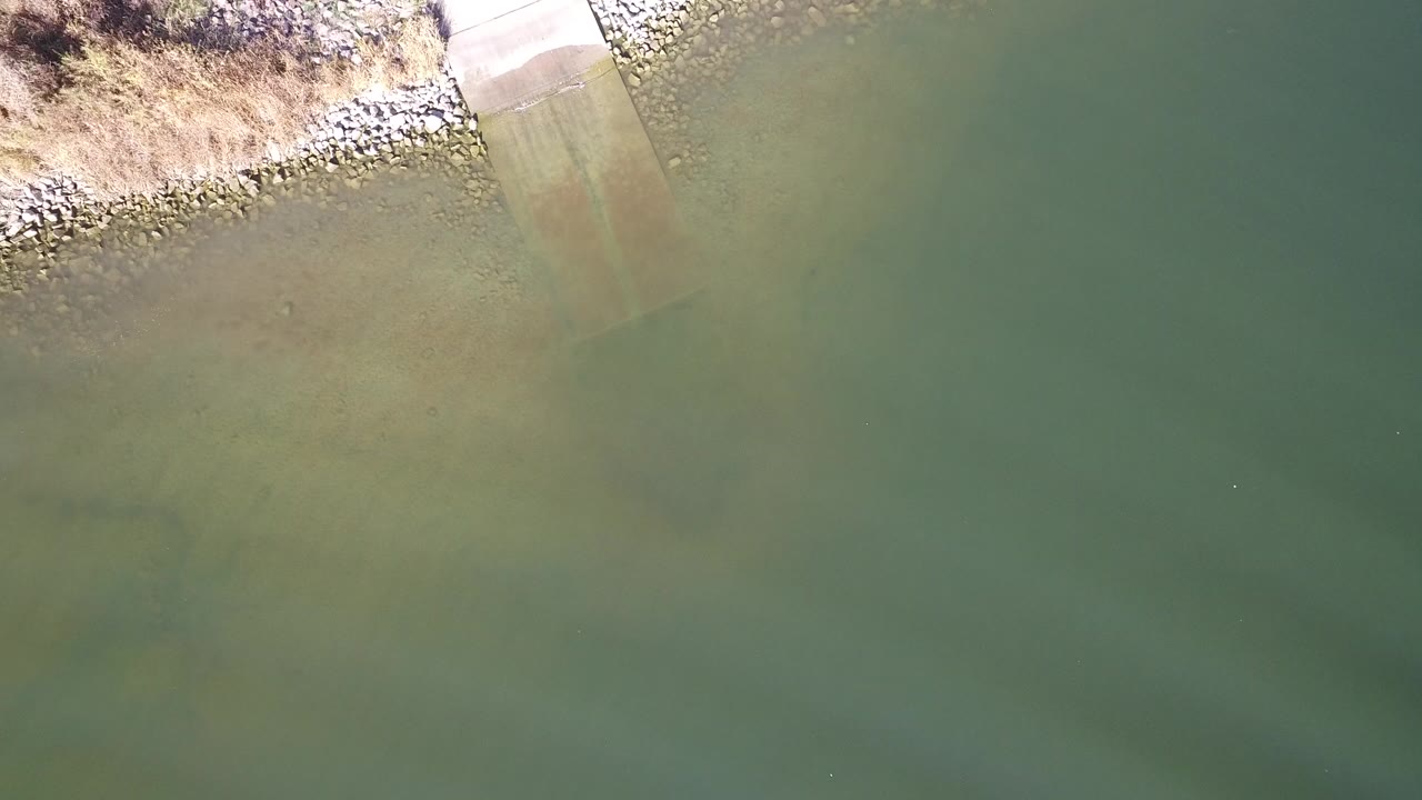 Overhead frame from Hidden Lake, Colorado showing green water, a pale ramp at the shore, riprap rocks, dry grass, and no visible people.