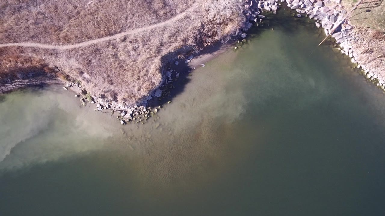 Overhead lake edge with green water, brown scrub terrain, rocks, and a narrow dirt path.