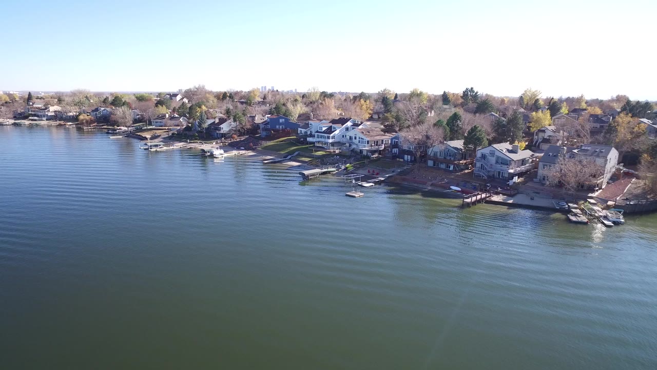 Opening aerial frame over Hidden Lake, Colorado, with blue-green water in front, shoreline houses and docks across the middle, bare trees behind them, and no visible people.