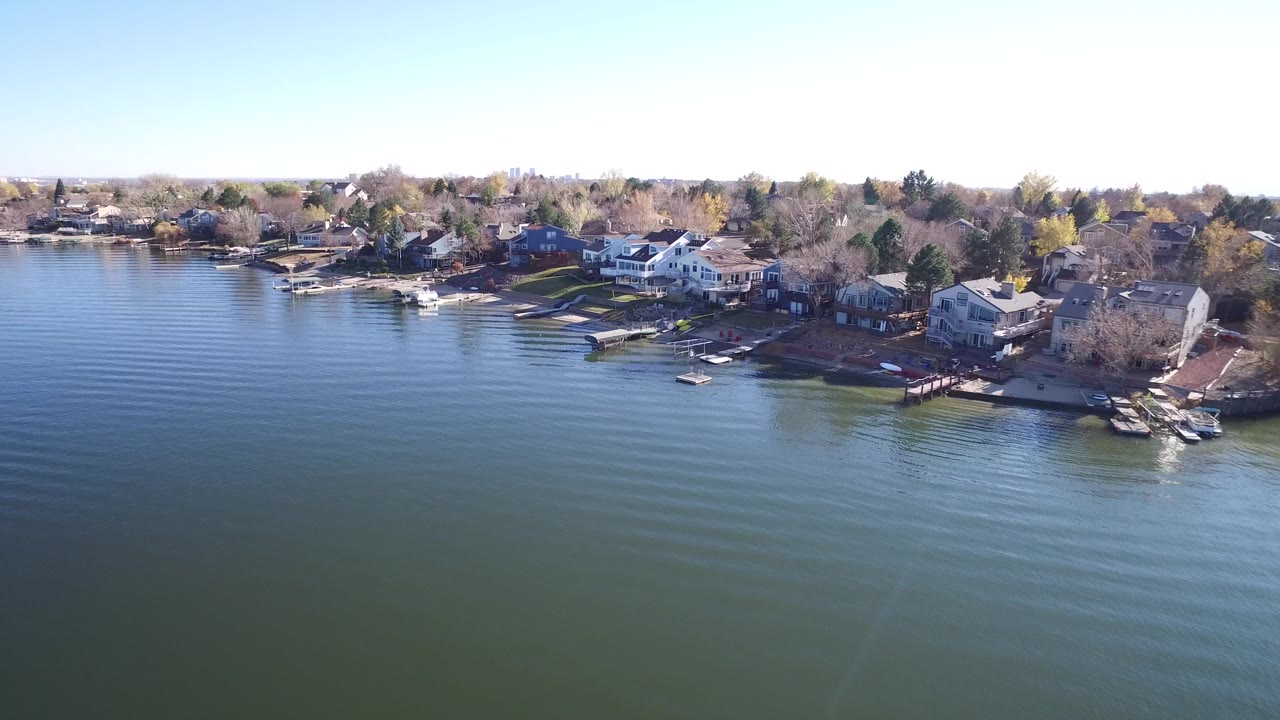 Early aerial frame of Hidden Lake, Colorado, where calm ripples cover the foreground and a row of waterfront homes, docks, lawns, and trees sits under a clear sky, with no visible people.