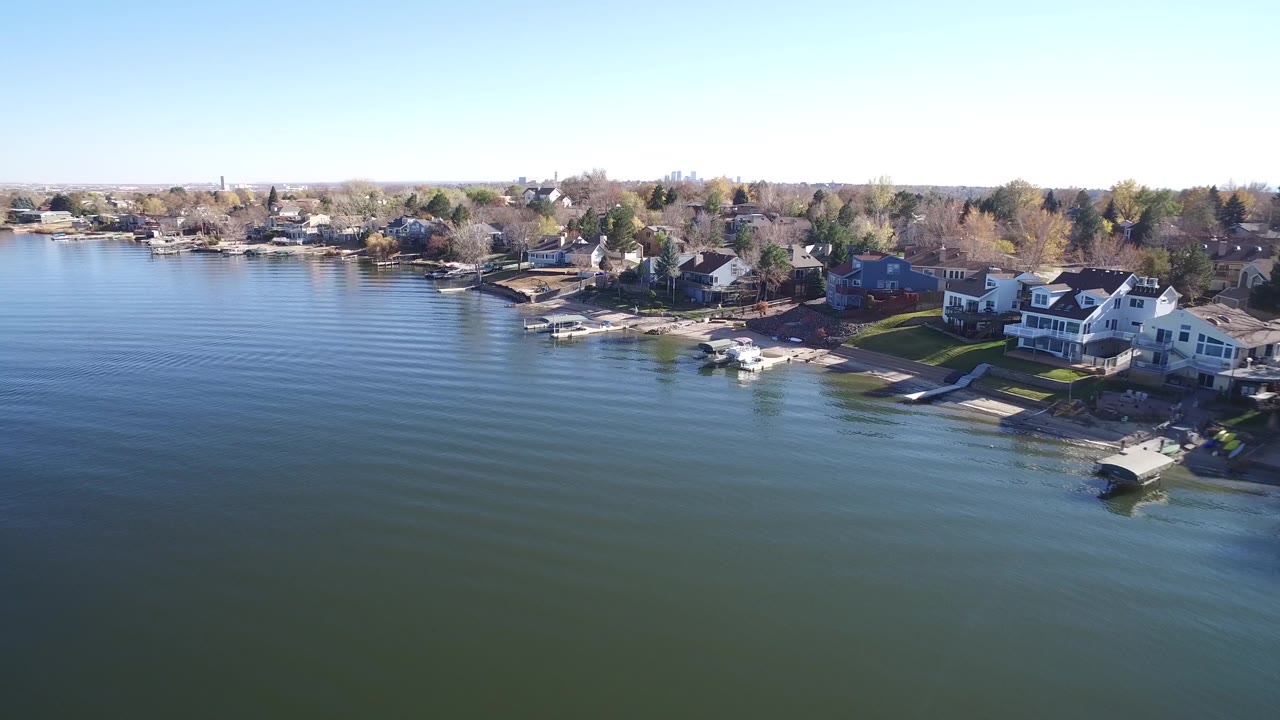 Wide aerial frame of Hidden Lake with rippled water in front, a row of homes and docks along the shore, and bright sky above, with no visible people.