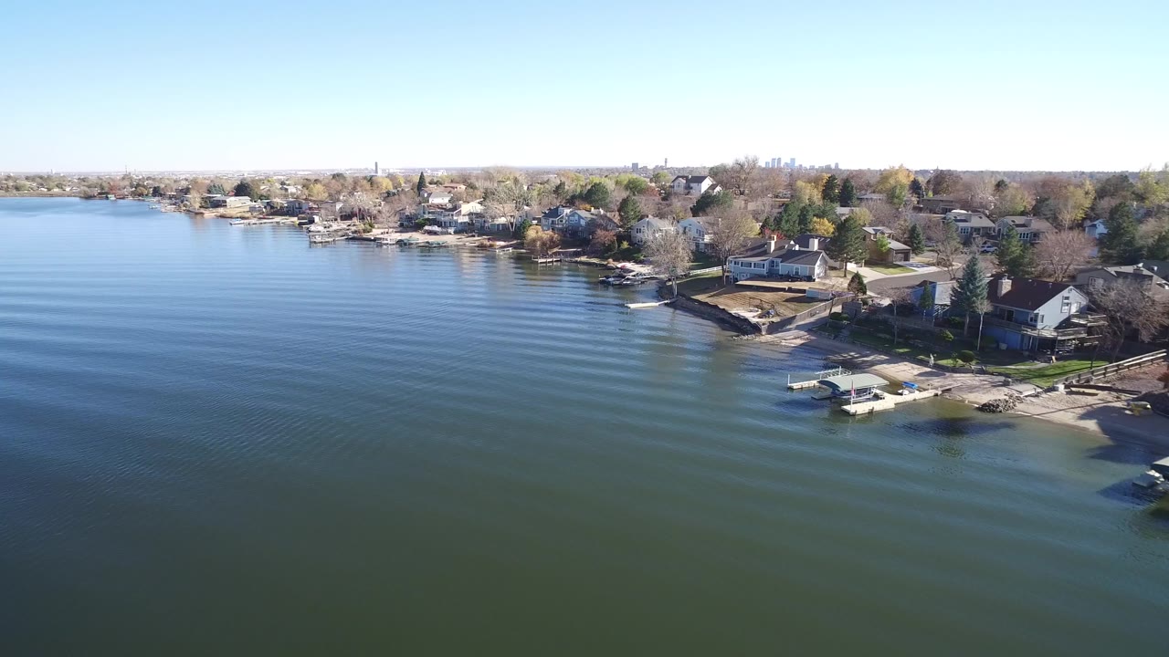 Early aerial frame over Hidden Lake, Colorado, with broad rippled water in the foreground, clustered lakeside homes, docks along the bank, and no visible people.