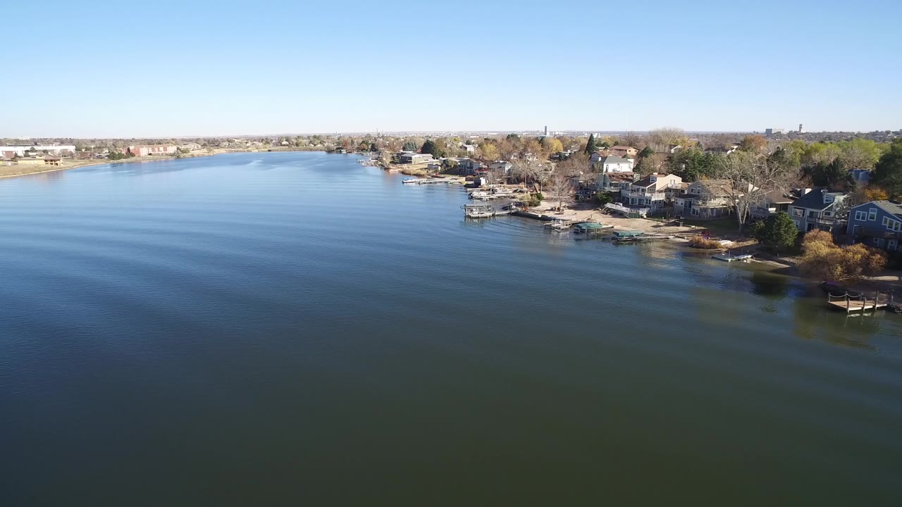 Opening aerial frame over Hidden Lake, Colorado, with blue-green water in the foreground, homes and docks along the right bank, low development on the far shore, and no visible people.