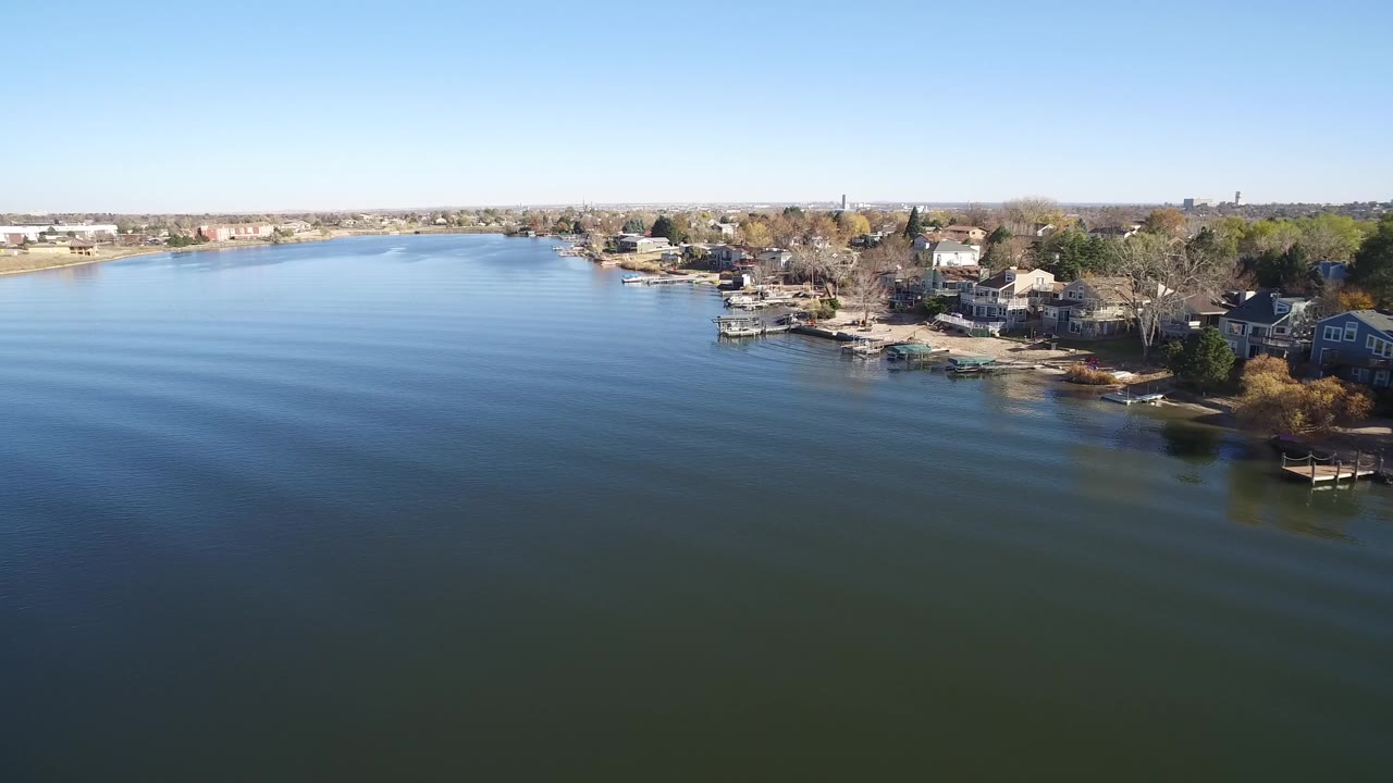 Hidden Lake, Colorado appears from a high aerial angle, with rippled water across most of the frame, a cluster of shoreline houses at right, distant buildings at left, and no visible people.