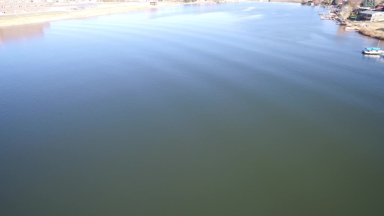 Elevated view over Hidden Lake, Colorado with blue-green water filling the frame, bright shoreline and small lakeside structures along the top edge, and no visible people.