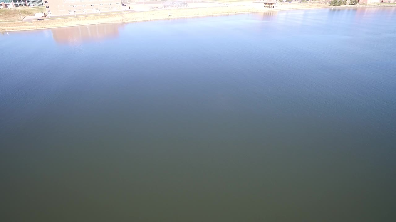 Aerial view over blue-green lake water with a bright shoreline at the top edge.