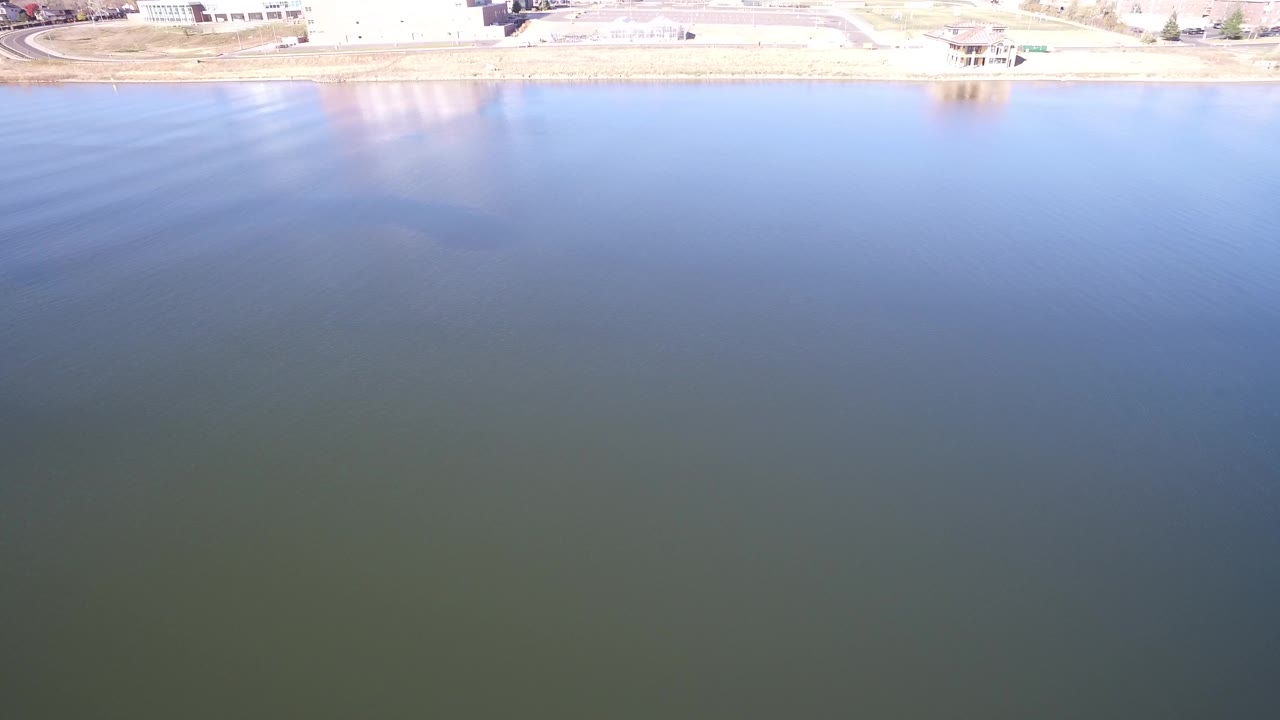 A high aerial frame over Hidden Lake, Colorado shows blue-green lagoon water filling the view, a pale sandy shoreline and road along the top edge, and no visible people.